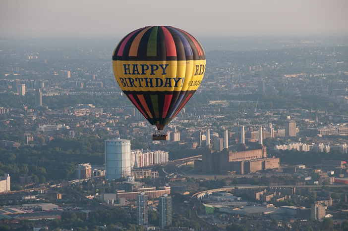 Happy Birthday Balloon Flight over London
Looking north over London with the iconic Battersea Power Station just to the left of our hot air balloon. The large green area just below the words Happy Birthday on the balloon is Hyde Park and the Serpentine.
Flights over London are very weather dependant, more so than a balloon flight over the home counties because as well as meeting the normal criteria for a hot air balloon flight which are light winds, no rain and good visibility, for a hot air balloon flight over London we are limited in the wind directions we can fly on in order to keep out of Heathrow Airport. However if you can live with the cancellations, when you do fly all our passengers say it is worth it and you will join a small exclusive club of balloon flight passengers who can say they have flown over London.
So because of this it is not so easy to emulate the first balloon pilots who flew in England from sites in the city in the 1780&rsquo;s. They were free to fly in any direction and indeed some flights from London even crossed the channel into Europe! Charles Green was a celebrated gas balloonist who flew over 400 miles in 1836 from Vauxhall Gardens, just south of the River Thames and not far from the modern day MI5 building. He even flew through the night, landing not far from Nuremberg in Germany
Click Here to find out how much it costs to fly in this balloon from somewhere near you